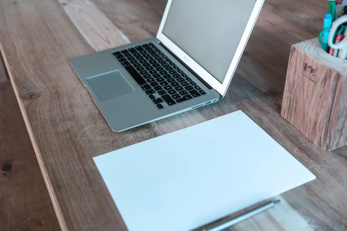 A laptop, blank paper and pen arranged on a wooden desk