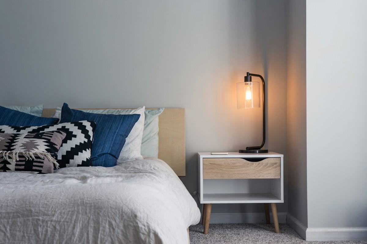 A bedside table and lit wall-mounted lamp beside patterned cushions