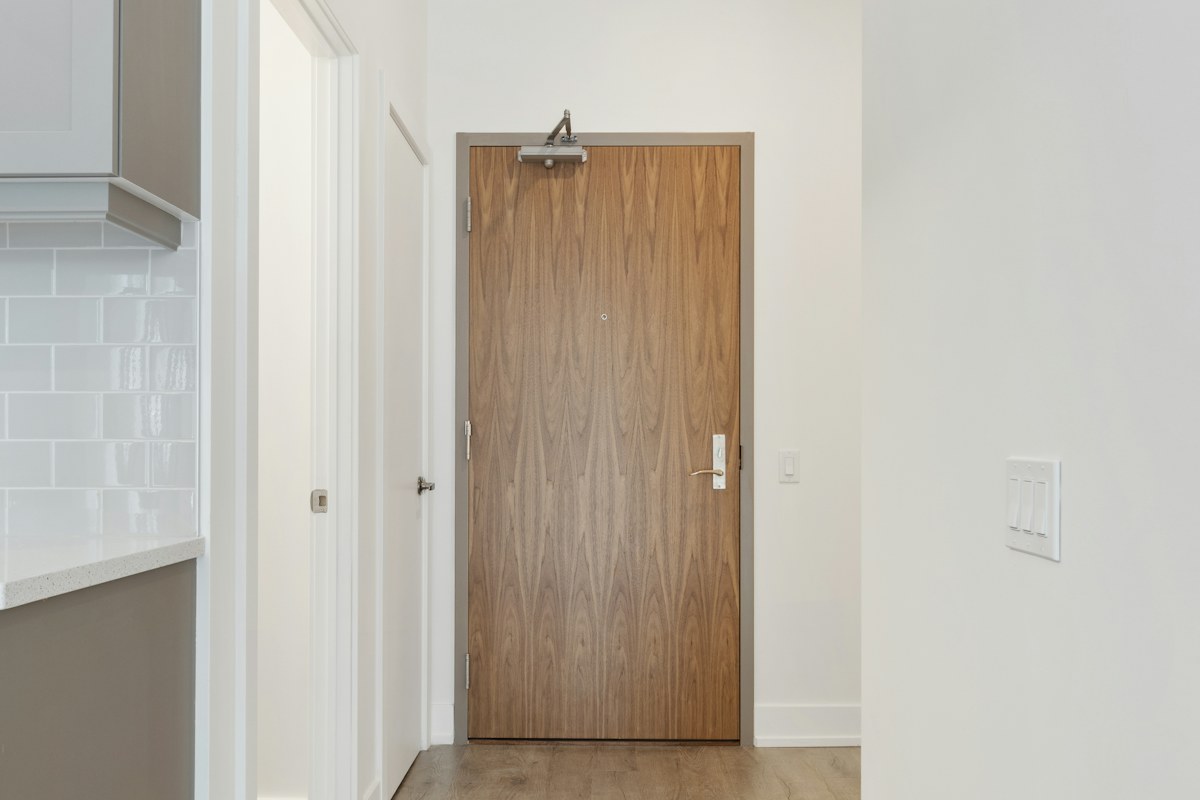 A wooden entrance door beneath a door closer in a pale hallway